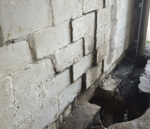 Cracked and damaged concrete block wall with missing sections beside a narrow, uneven trench. The scene suggests construction or repair work.
